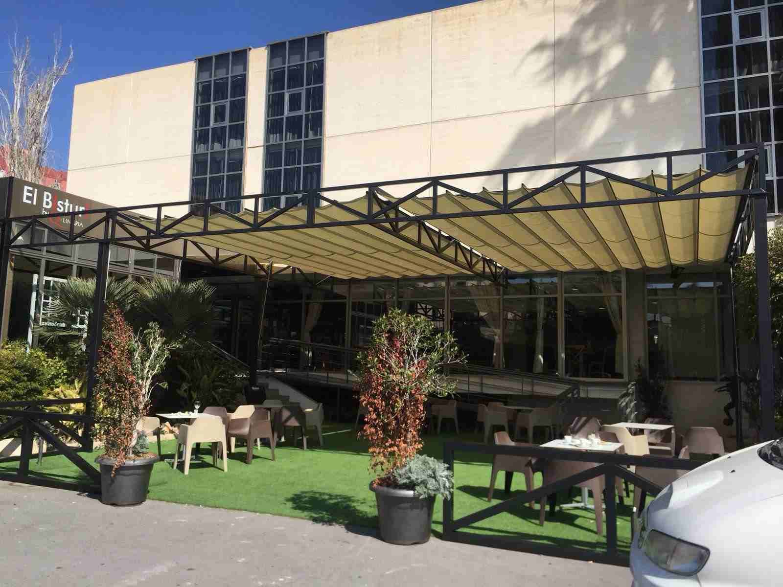 Un coche blanco está aparcado delante de un edificio con mesas y sillas bajo un dosel.