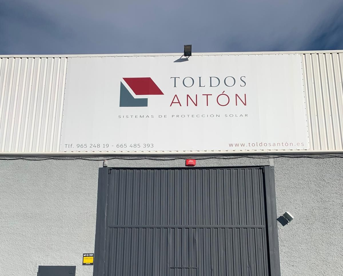Letrero de la empresa Toldos Antón en un edificio con una puerta gris cerrada. El logotipo incluye formas rojas y grises.