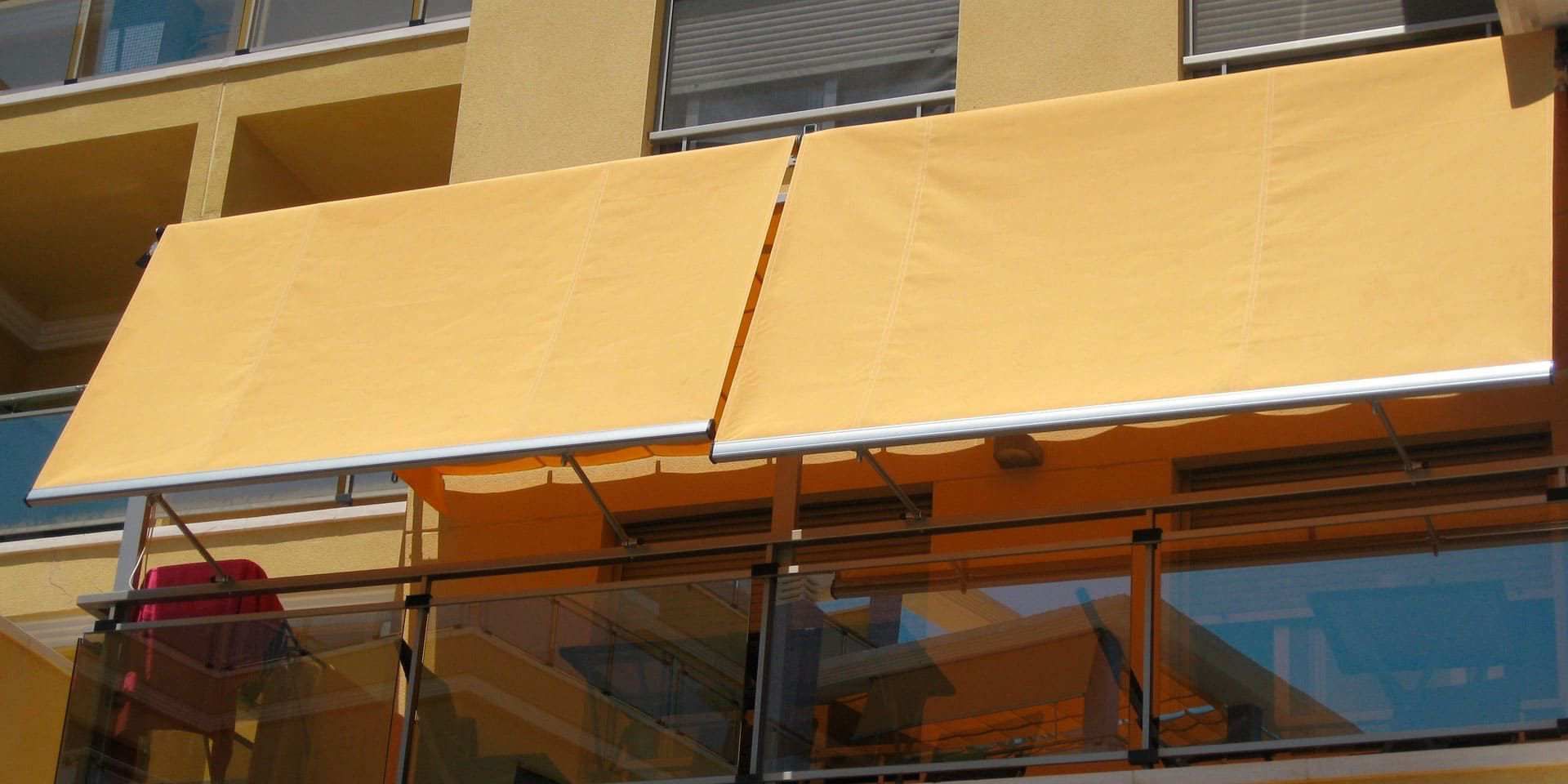 Dos toldos amarillos abiertos en un balcón con barandillas de cristal. Al fondo, el exterior del edificio.