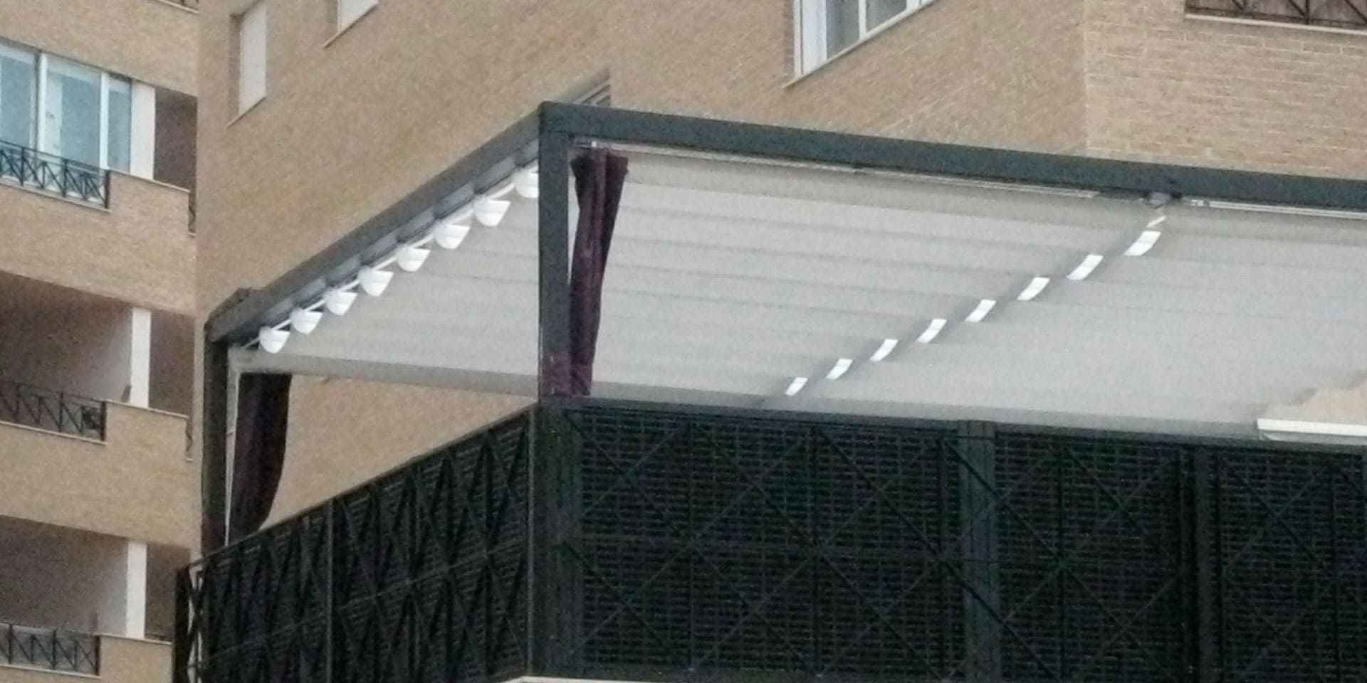 Patio exterior con techo retráctil blanco y valla oscura, junto a un edificio de ladrillo.