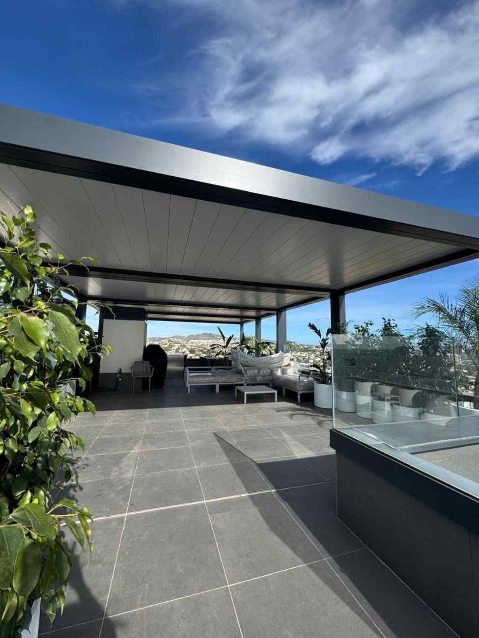 Patio en la azotea con pérgola moderna, muebles, plantas y vista a la ciudad bajo un cielo azul.