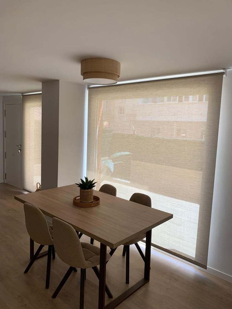 Comedor con mesa de madera, sillas y persianas beige. Una planta sobre la mesa.
