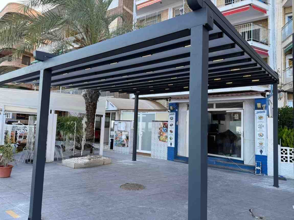 Pérgola negra sobre un pequeño negocio, con vista a la calle y una palmera al fondo.