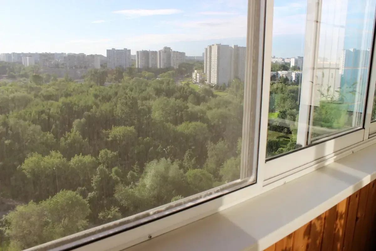 Vista desde una ventana con vistas a un bosque verde y a los edificios de la ciudad en un día soleado.