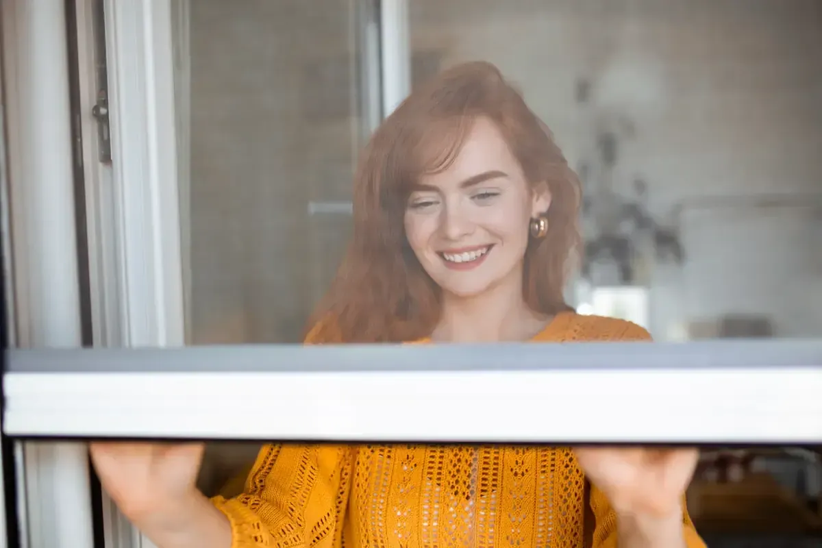 Mujer de cabello rojo sonriendo, mirando a través de una puerta mosquitera, vistiendo un suéter naranja.
