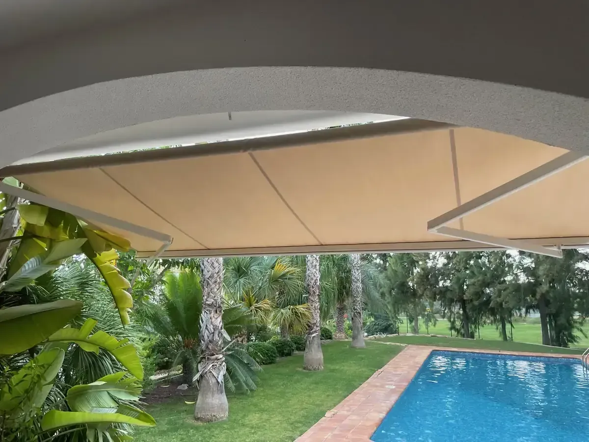 Un toldo retráctil beige da sombra a un área de piscina con una exuberante vegetación.