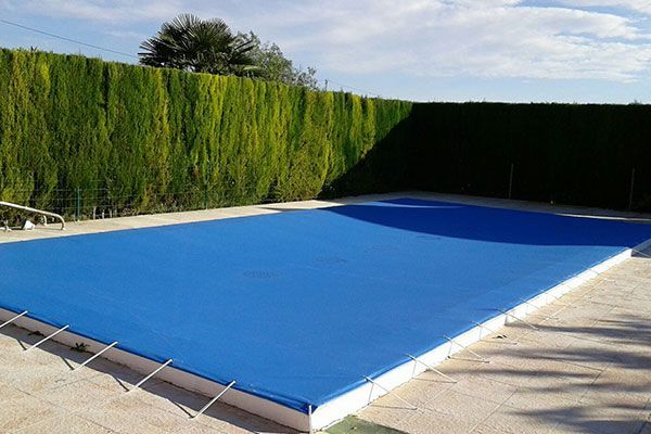 Cubierta azul sobre una piscina rectangular. Seto verde al fondo bajo un cielo azul.