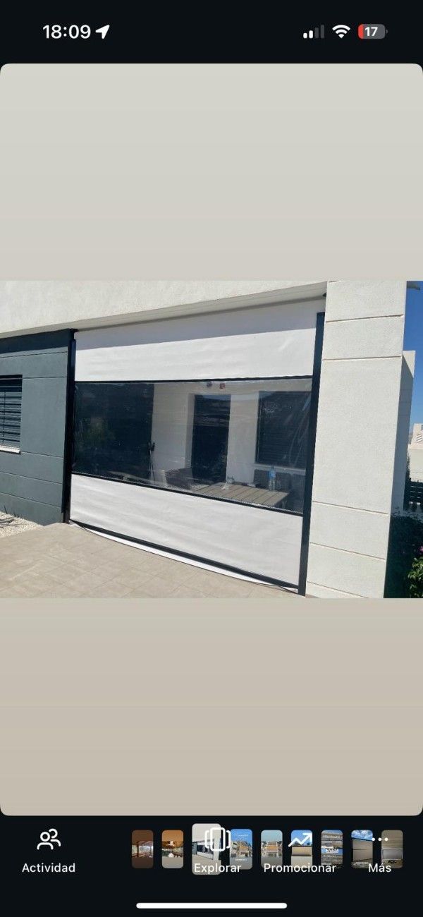 Persianas enrollables beige cubriendo parcialmente una ventana. Afuera, se ven edificios y árboles.