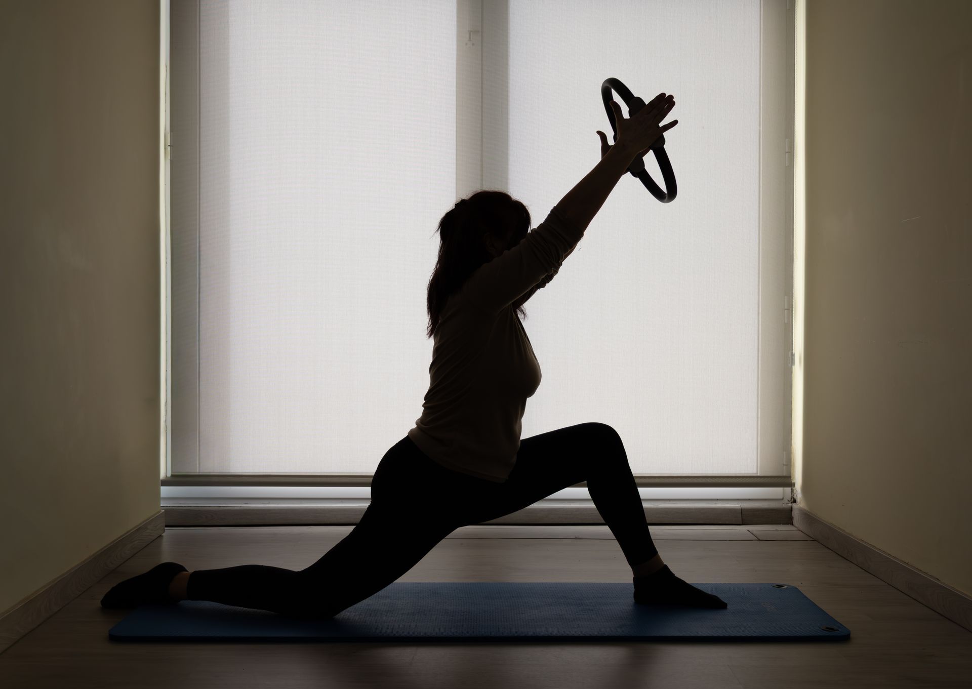 Silueta de una persona haciendo una zancada de yoga con los brazos levantados sobre una esterilla frente a una ventana luminosa.