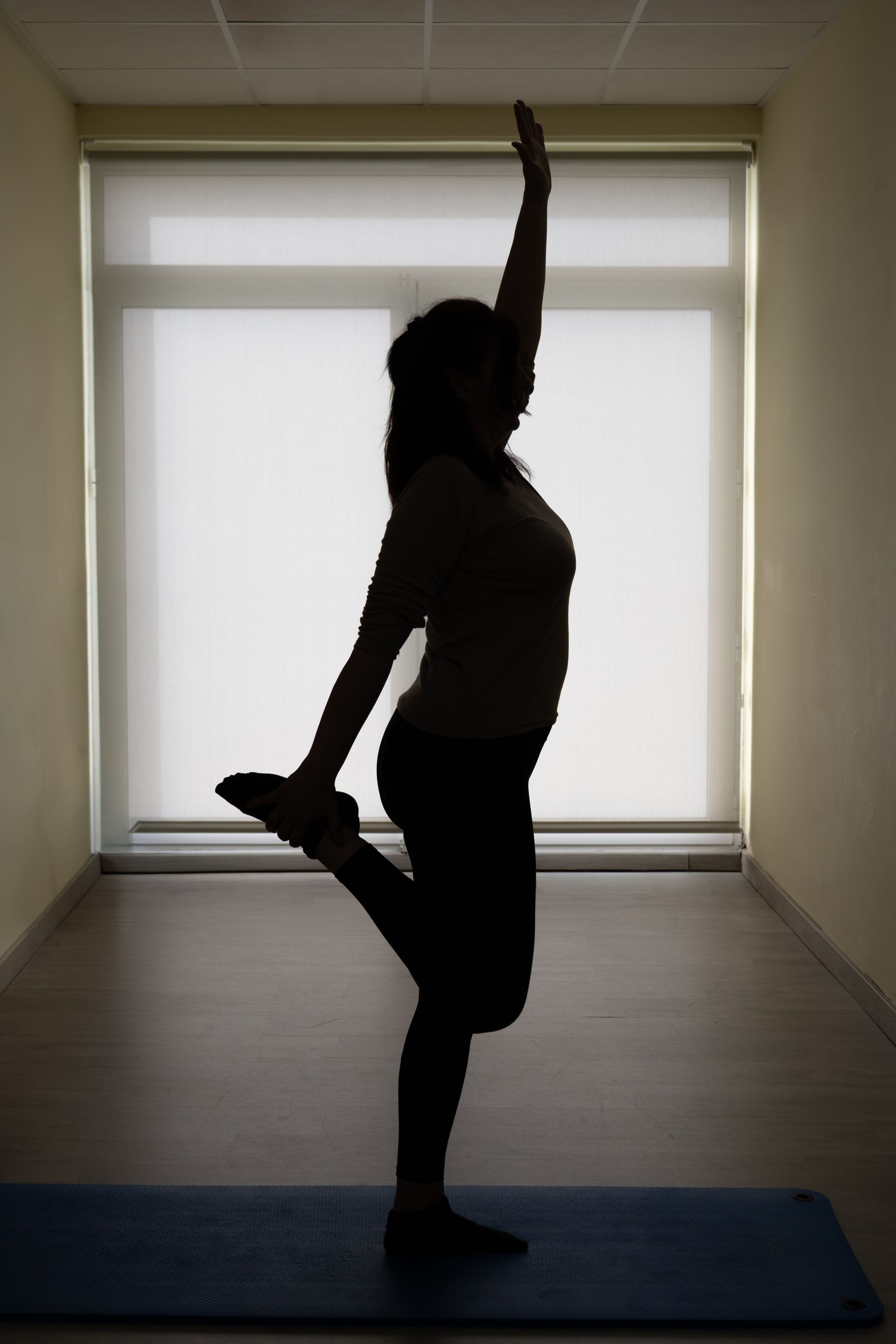 Silueta de una persona realizando una postura de yoga de pie sobre una esterilla en una habitación luminosa.