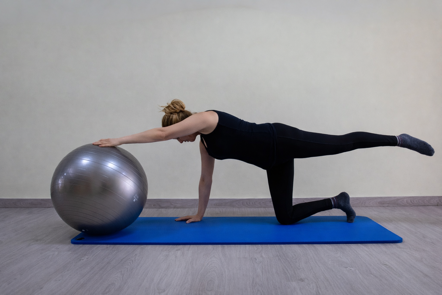 Una persona realiza un ejercicio de equilibrio sobre una colchoneta azul, con un brazo apoyado en una pelota de ejercicio plateada y una pierna levantada.