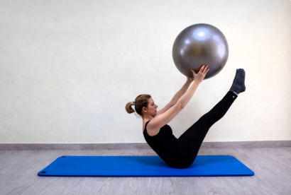 Persona realizando una postura en V sobre una colchoneta azul, sosteniendo una pelota de ejercicio plateada por encima de la cabeza.