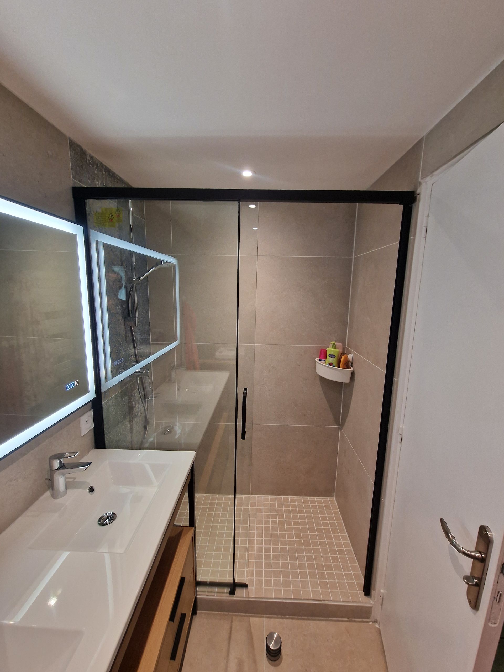 Salle de bain moderne avec douche, meuble-lavabo et porte de toilettes. Cabine de douche à cadre noir, carrelage gris, lavabo blanc et meuble en bois.