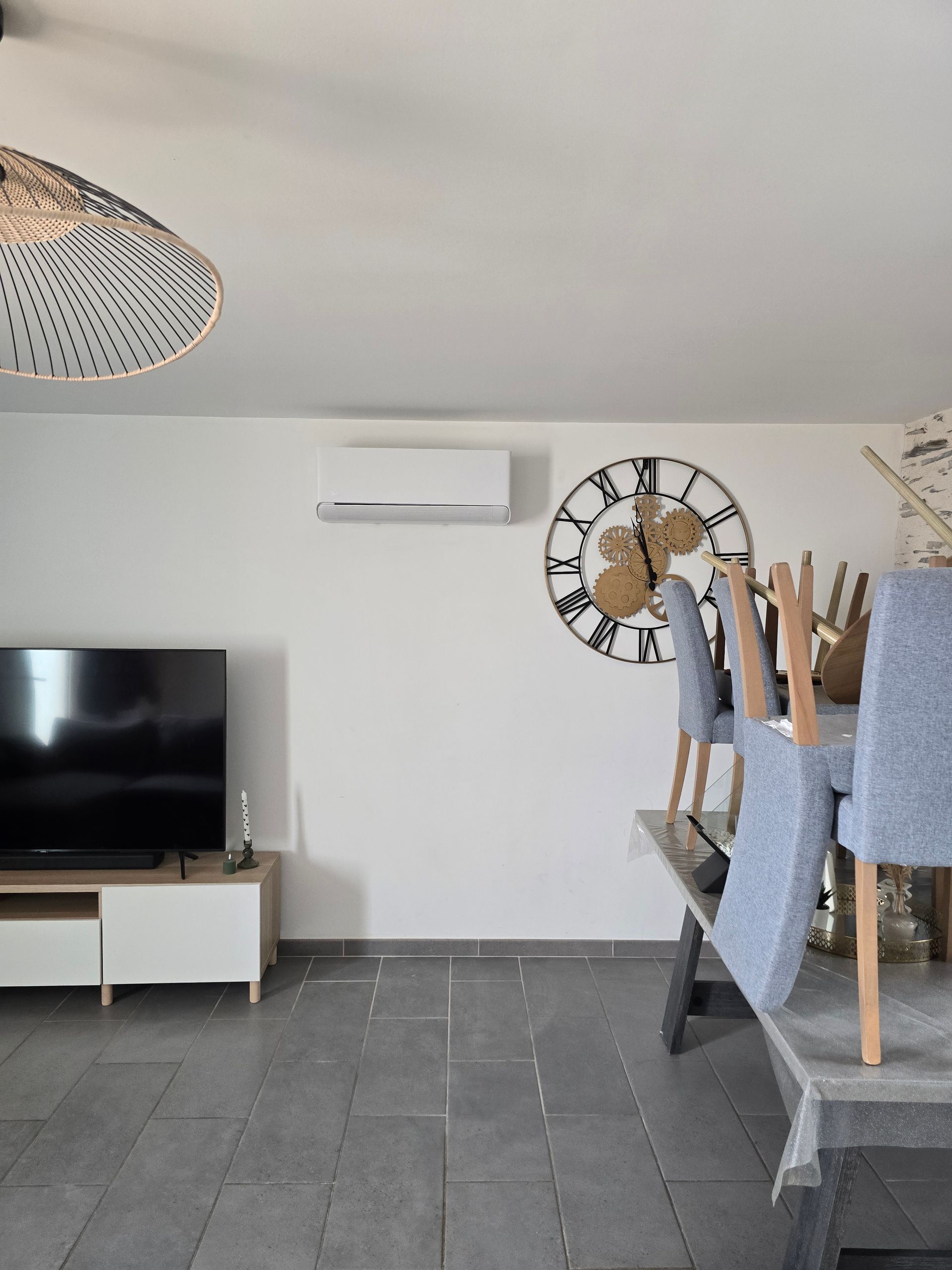 Salon avec télévision, climatiseur, grande horloge et chaises empilées sur une table.