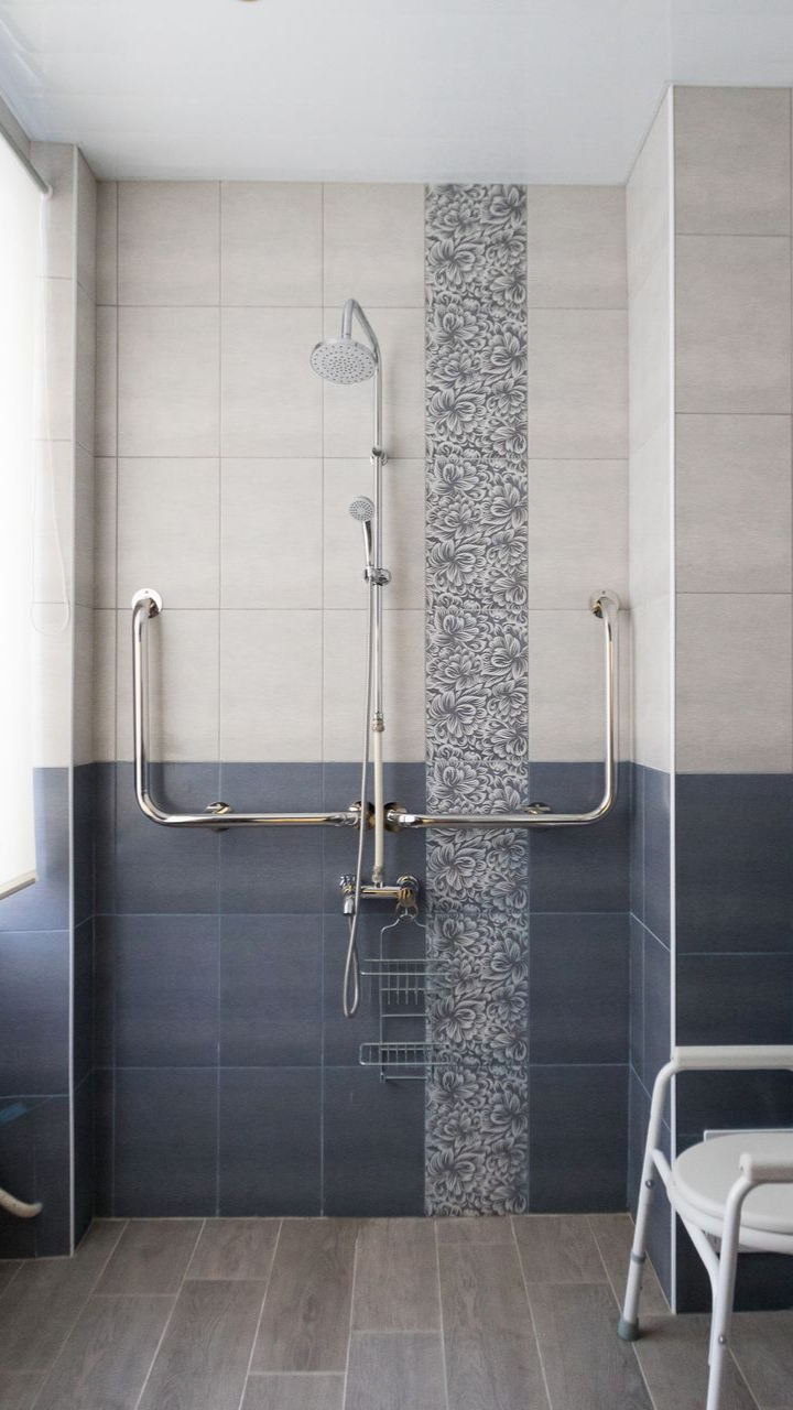 Douche accessible aux personnes à mobilité réduite avec mains courantes, carrelage gris et bleu, avec chaise de douche.