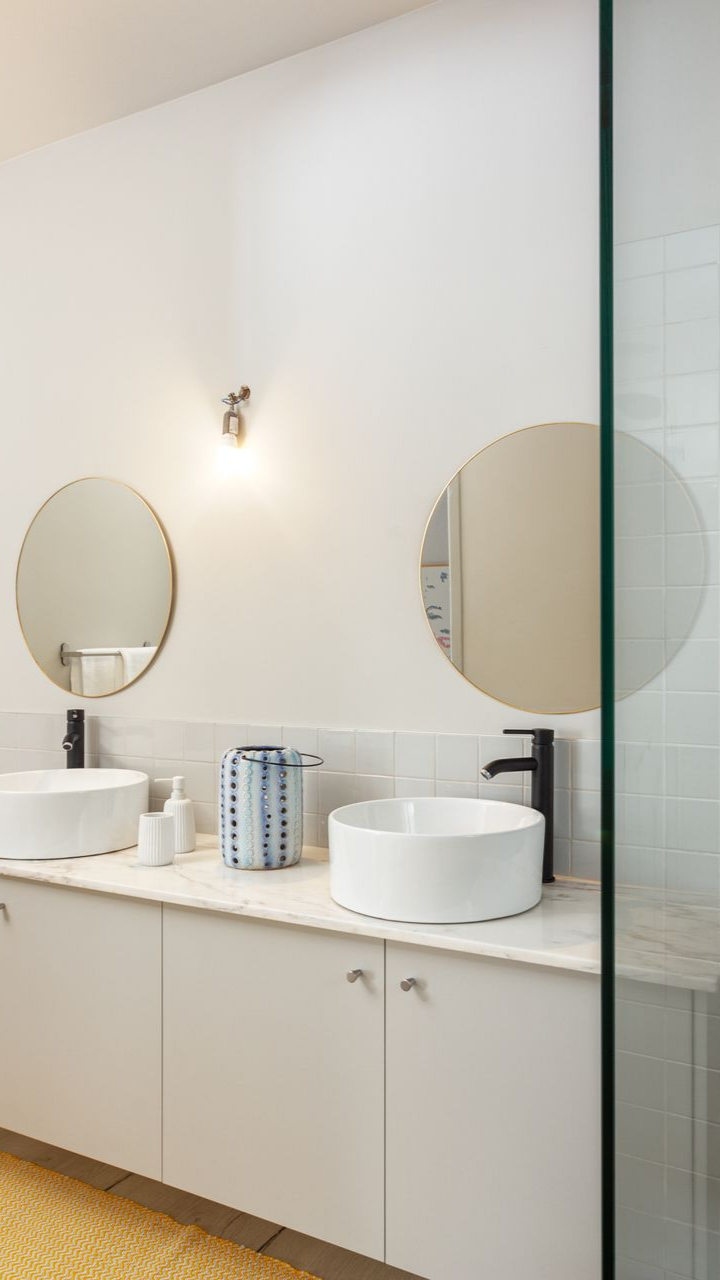 Salle de bains blanche avec deux lavabos, miroirs ronds, robinets noirs et une porte de douche en verre.