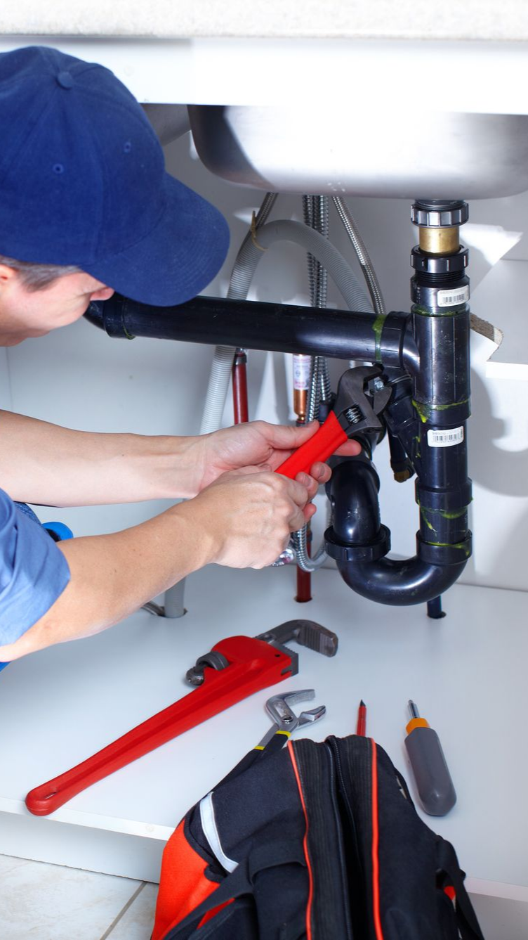 Plombier portant une casquette bleue utilisant une clé rouge pour réparer des tuyaux sous un évier.