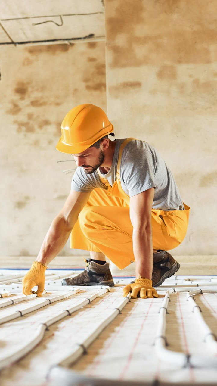 Ouvrier du bâtiment en combinaison jaune et casque de sécurité inspectant les tuyaux de chauffage par le sol.