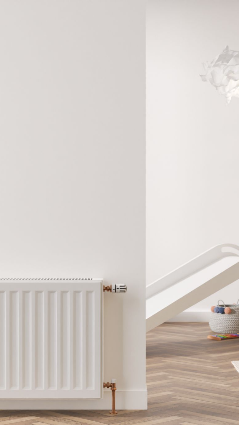 Radiateur blanc sur un mur blanc, avec tuyaux en cuivre. Parquet à chevrons, pièce lumineuse.