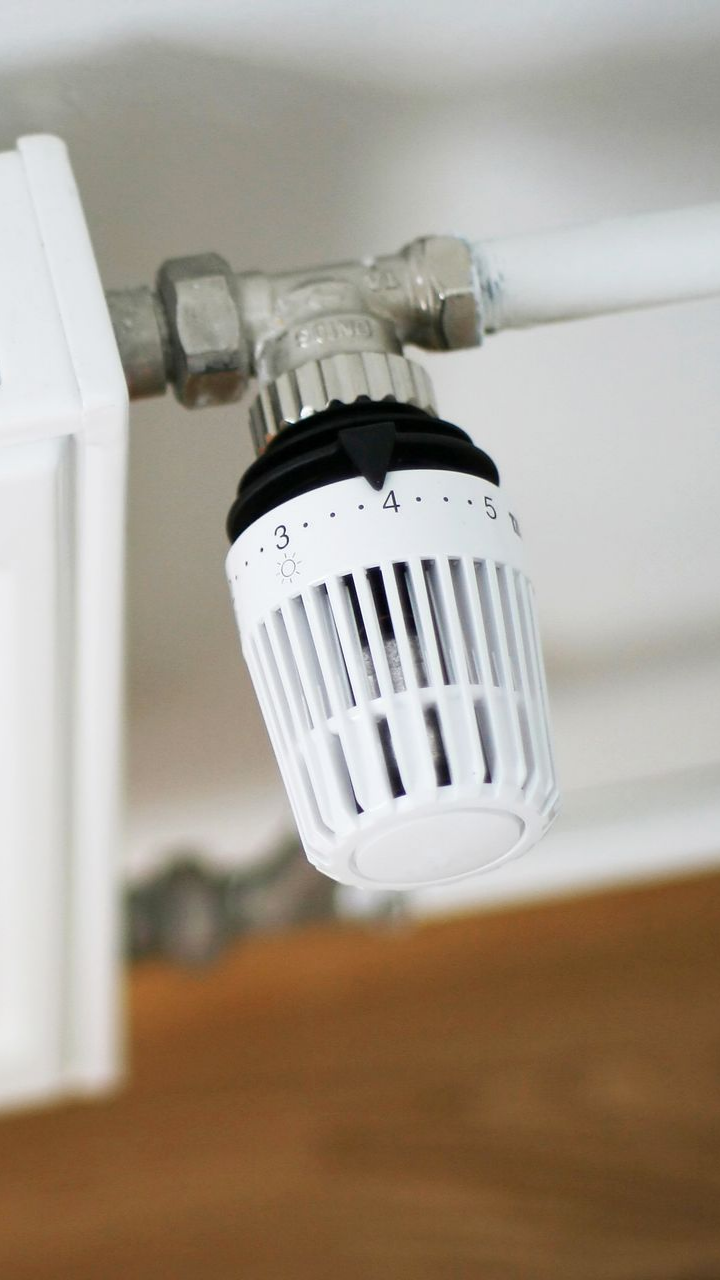 Robinet de radiateur avec cadran numéroté, monté sur tuyau blanc et radiateur, près d'un plancher en bois.