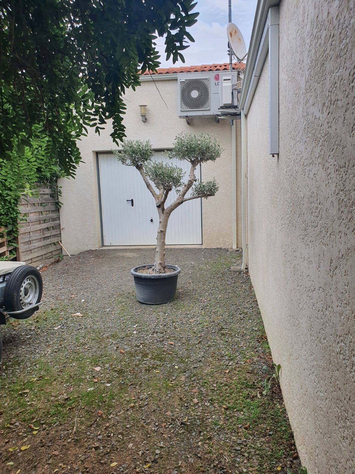 Unité de climatisation montée sur un extérieur de bâtiment de couleur claire avec un toit en tuiles rouges, à proximité d'un petit jardin.