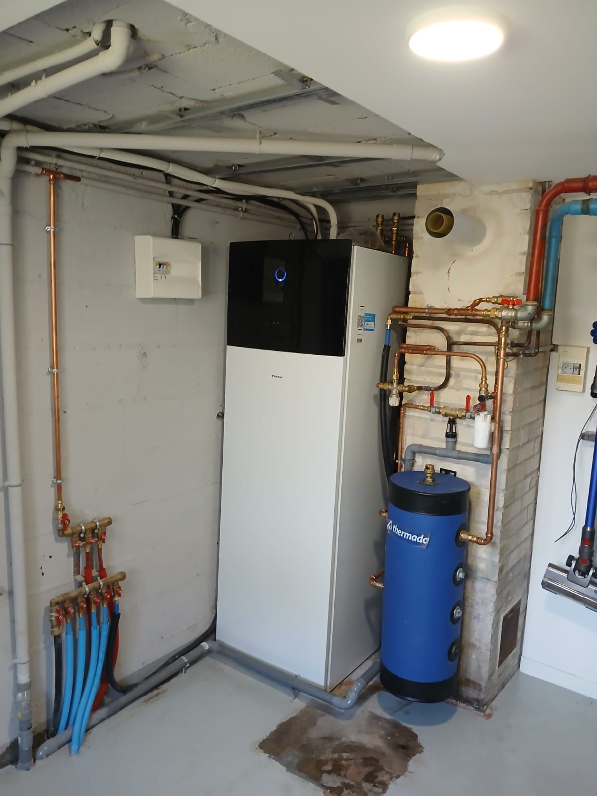 Installation d'une PAC air-eau dans un sous-sol. Unité blanche, réservoir bleu, tuyaux en cuivre, murs gris.
