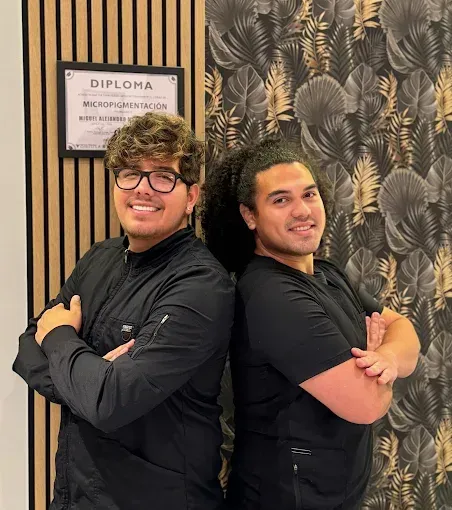 Dos hombres, con los brazos cruzados, sonríen a la cámara. Uno tiene el pelo rizado y gafas. 