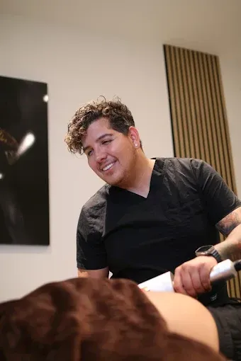 Un hombre vestido de negro sonríe mientras sostiene un dispositivo sobre la espalda de alguien: un tratamiento estético.