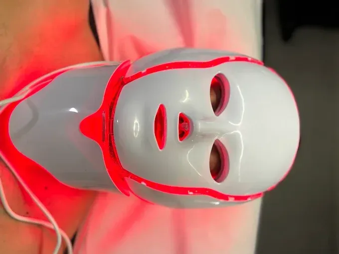 Máscara de terapia de luz LED blanca en la cara de una persona, emitiendo luz roja.