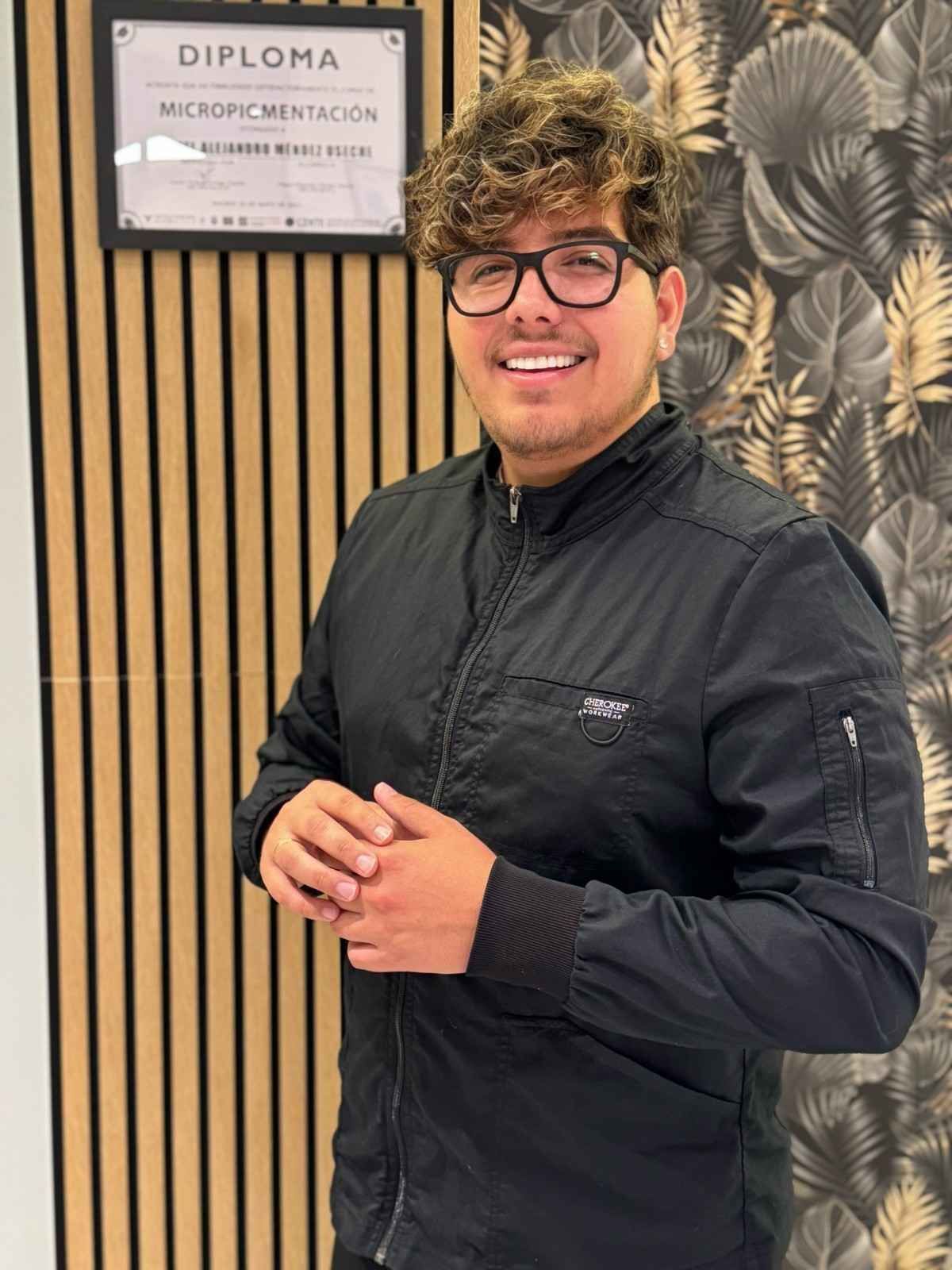 Hombre con gafas y chaqueta negra sonríe cerca de un diploma de micropigmentación.