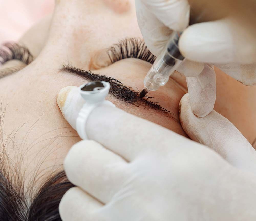 Un profesional utiliza un lápiz de tatuaje para realizar la técnica de microblading en la ceja de una persona, con la tinta contenida en un recipiente con forma de anillo.