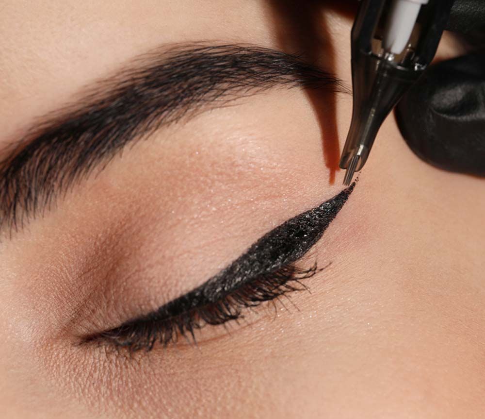 Una mano enguantada utiliza una herramienta especializada para tatuar para aplicar delineador de ojos negro permanente en un párpado.