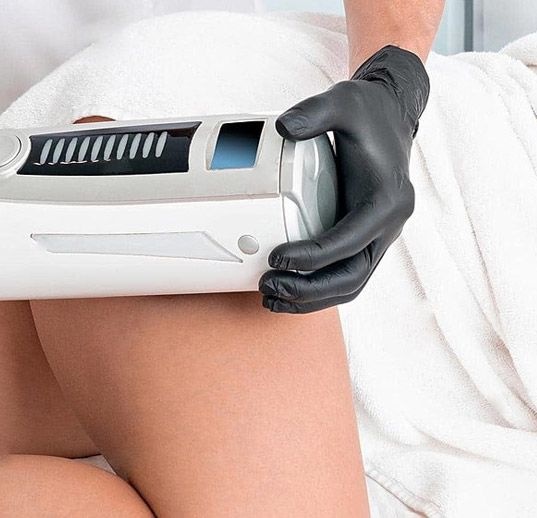 En un entorno clínico, una persona con guantes negros sostiene un dispositivo médico cosmético contra la parte superior del muslo de un paciente.