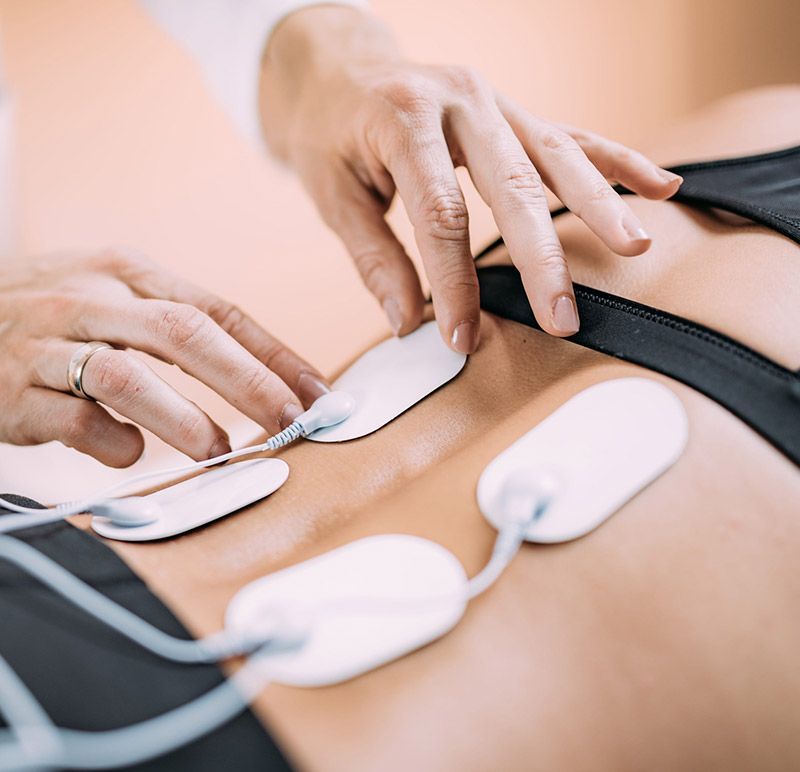 Manos colocando electrodos adhesivos de estimulación eléctrica en la parte baja de la espalda de una persona para un tratamiento de fisioterapia.