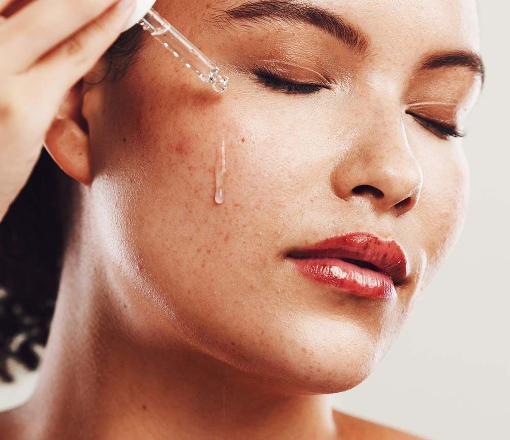 Una persona se aplica un sérum transparente en la mejilla con un cuentagotas de cristal, con los ojos cerrados y expresión tranquila.