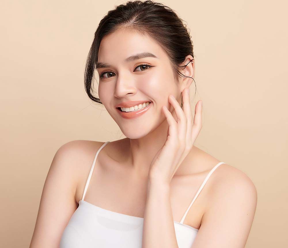 Una persona sonriente con una piel radiante y tersa se toca suavemente la mejilla sobre un fondo beige neutro y uniforme.