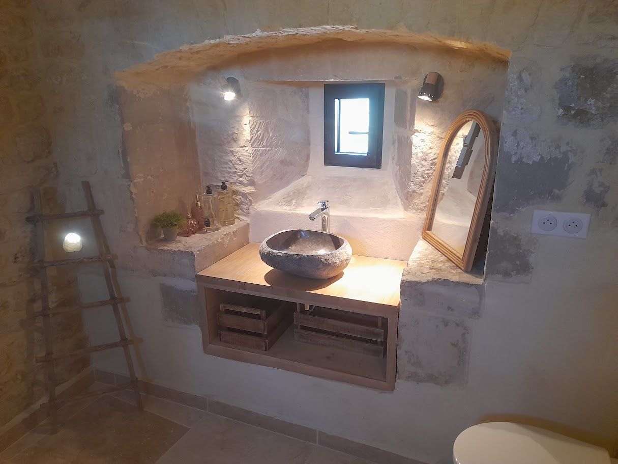 Une salle de bains aux murs de pierre, avec un lavabo vasque en pierre naturelle sur une étagère en bois, une petite fenêtre et un porte-serviettes incliné.