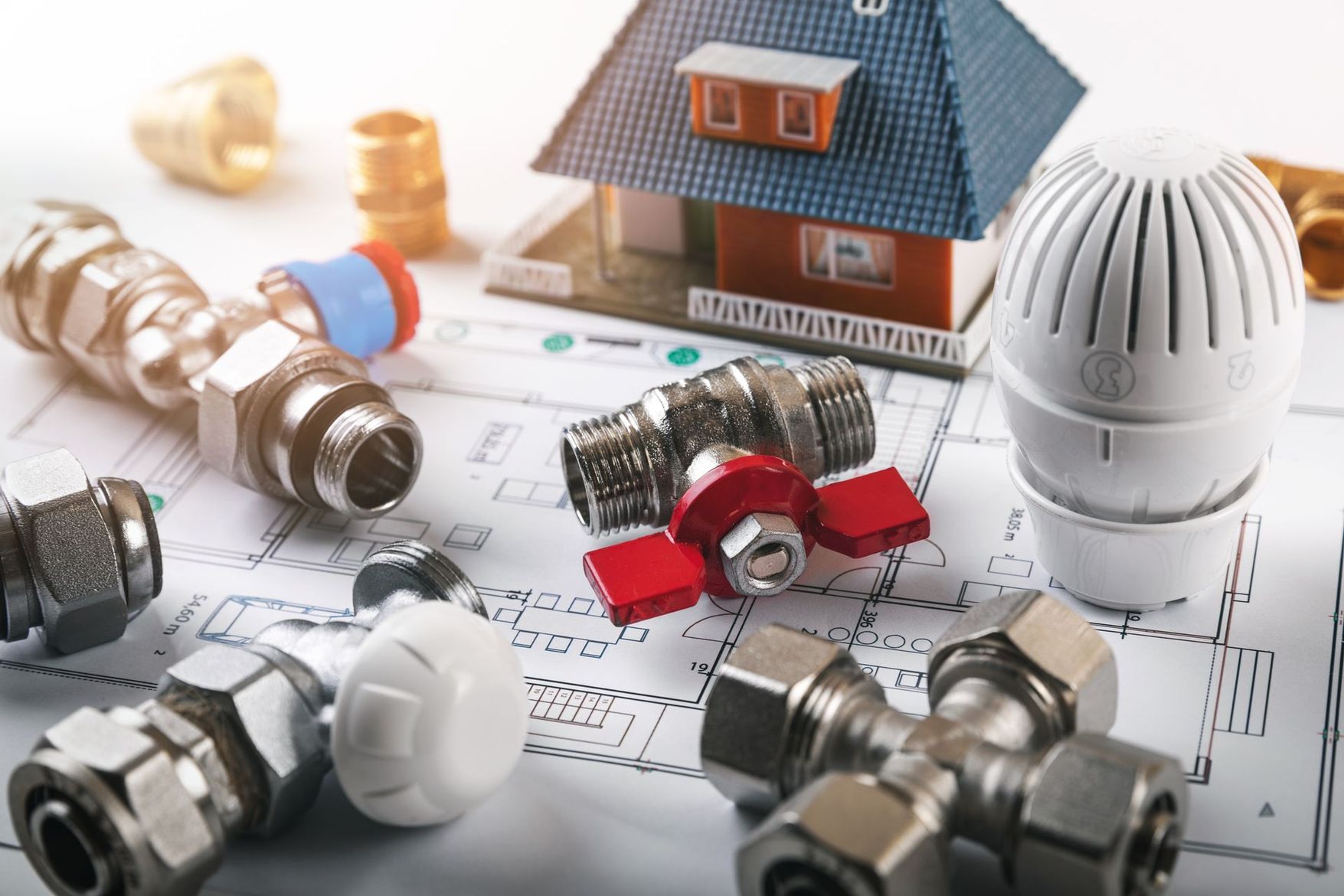 Raccords de plomberie, vannes et thermostat de radiateur disposés sur des plans architecturaux à côté d'une maquette de maison.