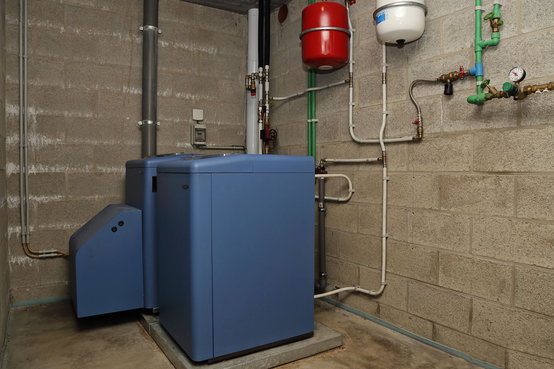 Une chaudière bleue dans un sous-sol aux murs en béton apparents, avec tuyauterie et vases d'expansion rouges et blancs.