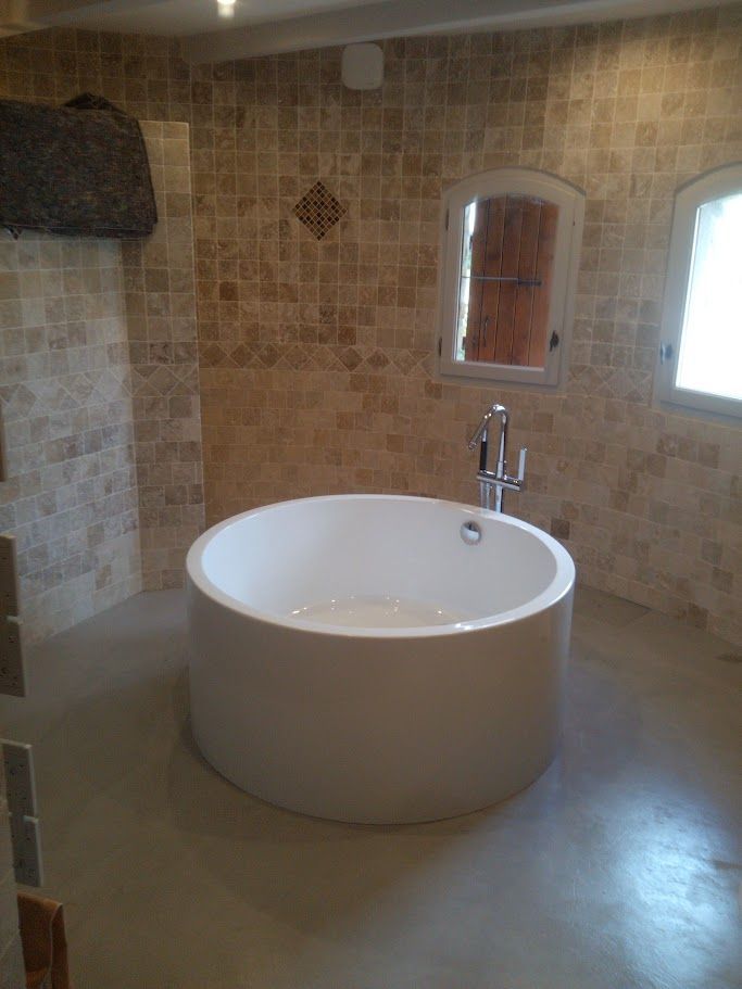 Une baignoire blanche ronde sur pieds trône dans une salle de bains aux murs carrelés de beige texturé et au sol lisse.