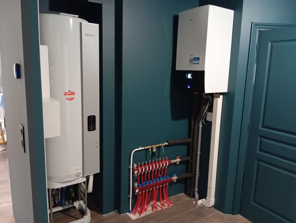 Une chaudière et un chauffe-eau muraux blancs sont installés contre un mur turquoise, à côté d'un collecteur de chauffage avec des tuyaux rouges et bleus.