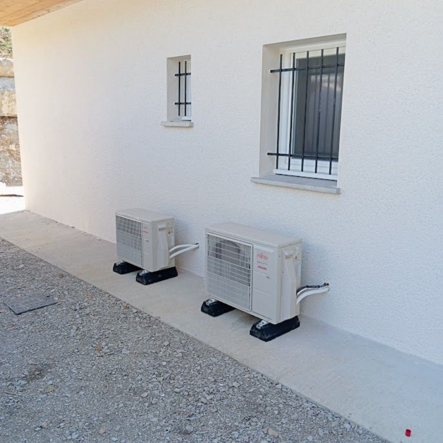 Deux unités extérieures de climatisation sont installées sur des dalles de béton contre un mur extérieur blanc percé de deux fenêtres.