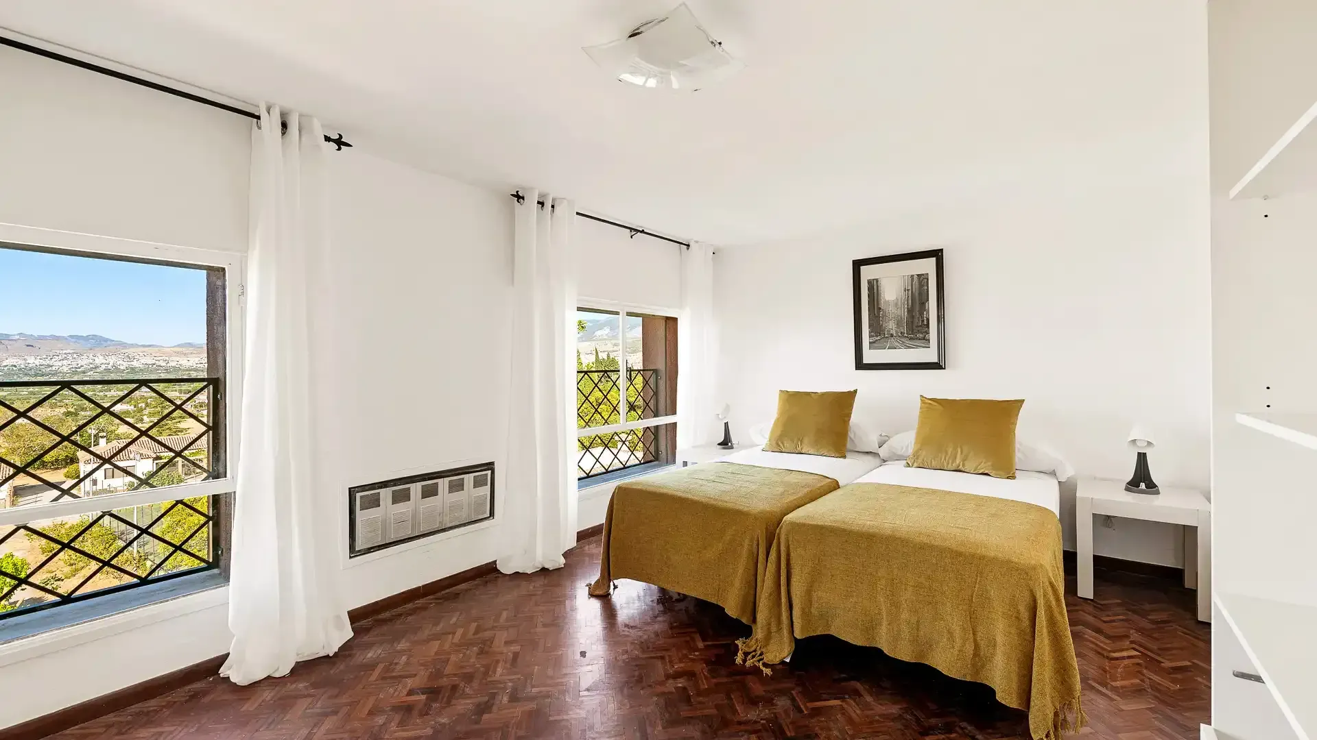 Habitación luminosa con paredes blancas, ropa de cama color mostaza, suelo de madera y grandes ventanales con cortinas.