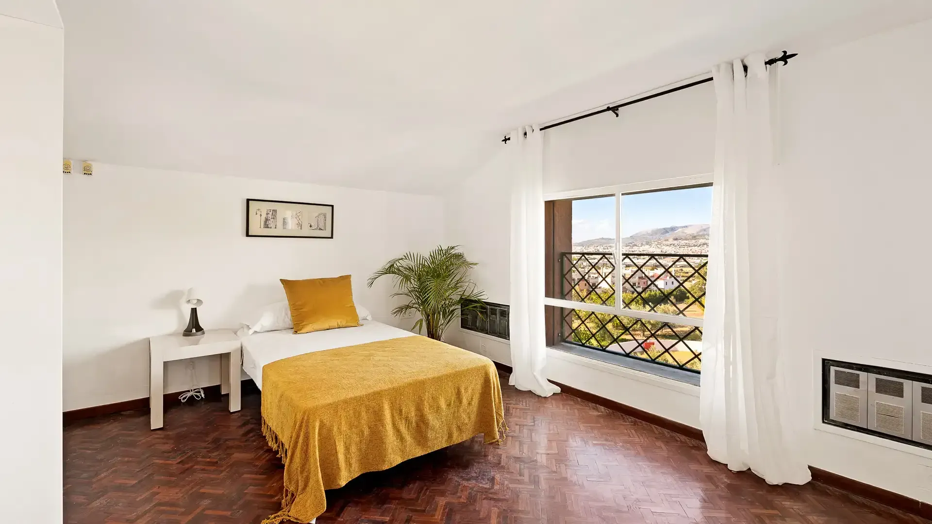 Habitación luminosa con paredes blancas, colcha amarilla, mesita de noche, planta y gran ventanal con vistas al exterior.