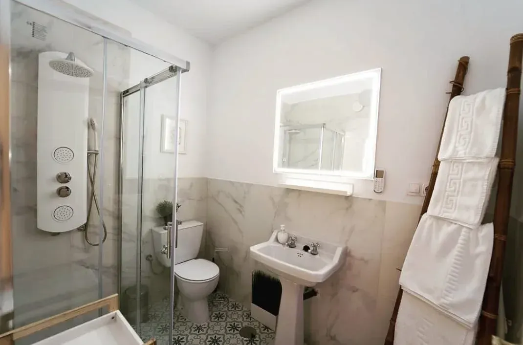 Baño blanco luminoso con ducha de cristal, inodoro, lavabo y toallero a la derecha.