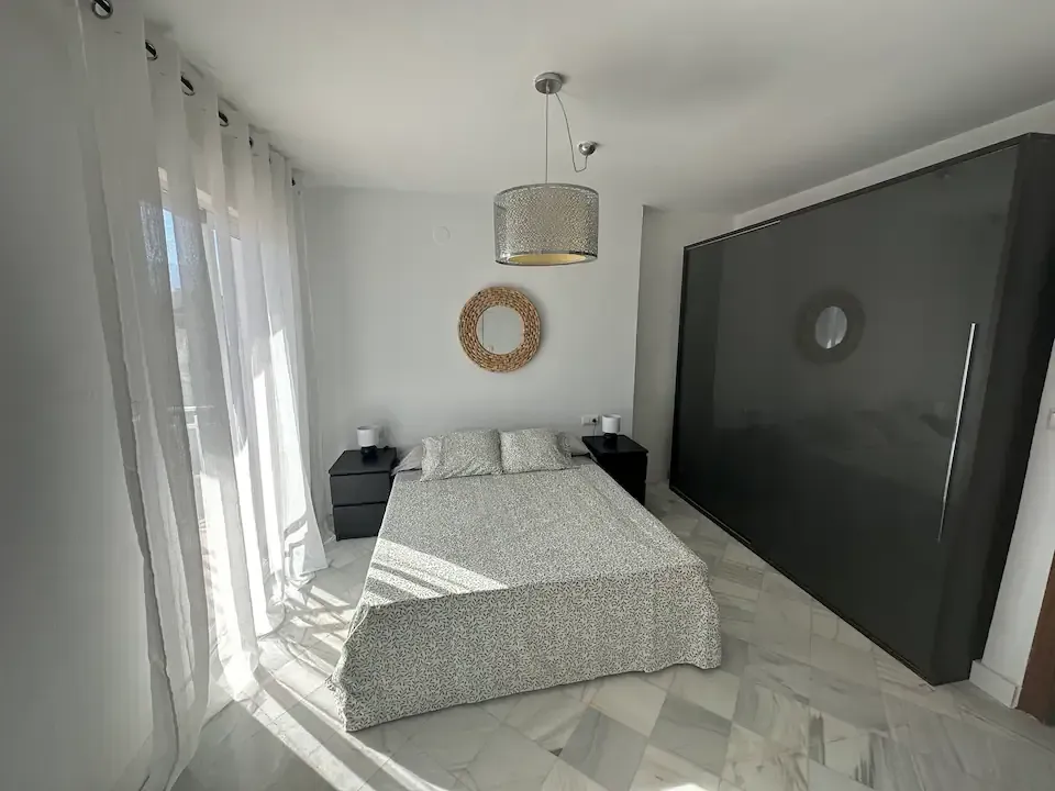 Habitación luminosa con cama gris, cortinas blancas, pared con espejo y suelo de espiga iluminado por la luz del sol.