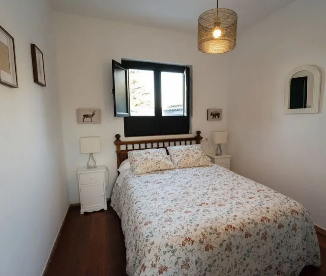 Habitación pequeña de color blanco con cama doble, ventana con marco negro y lámpara de techo de mimbre.