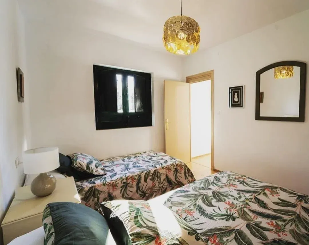 Habitación luminosa con dos camas estampadas, paredes blancas, cuadros negros en las paredes y una lámpara colgante.