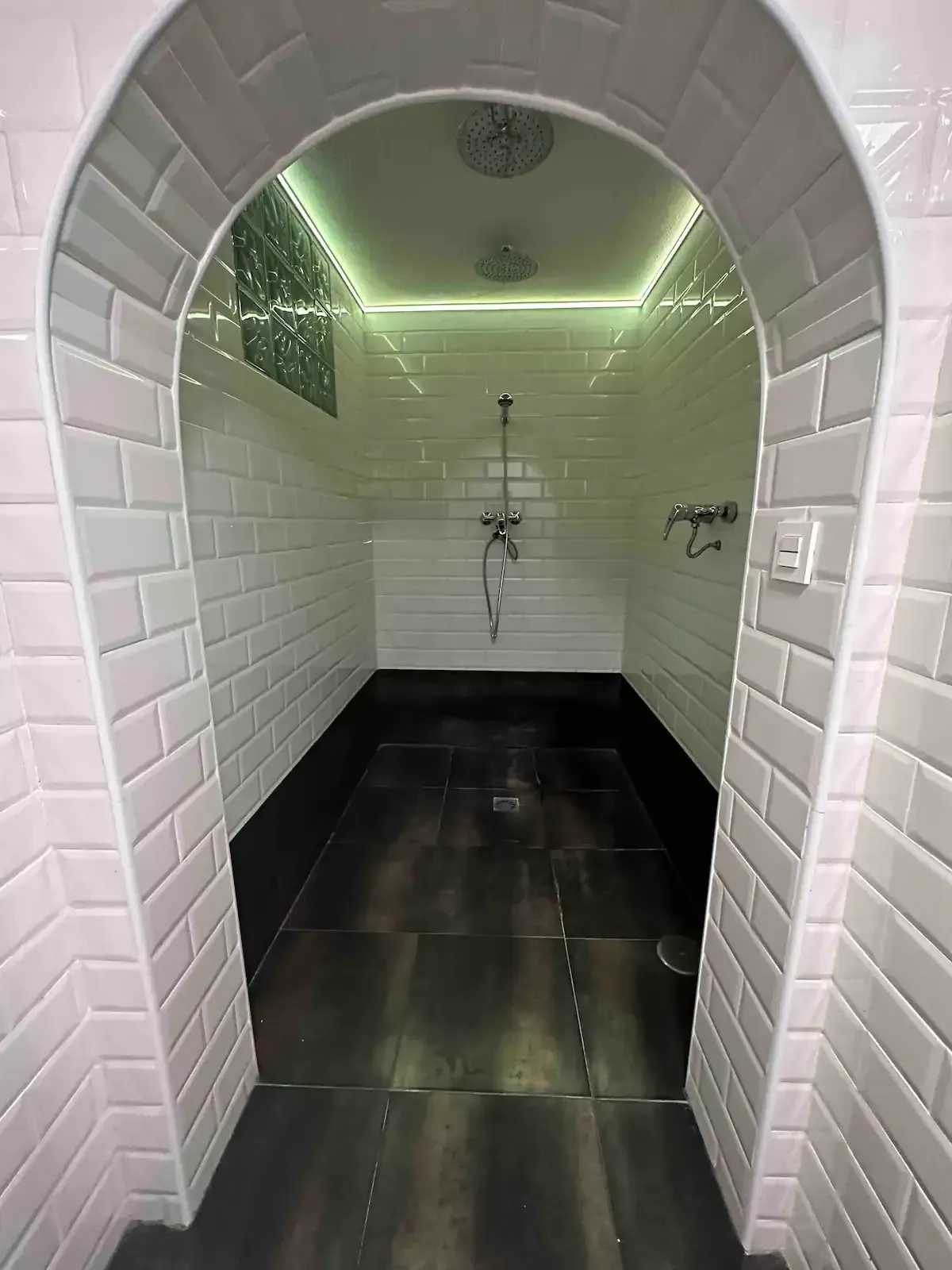 Cuarto de ducha abovedado con azulejos blancos, suelo de baldosas oscuras y tira de iluminación LED verde bajo el techo.