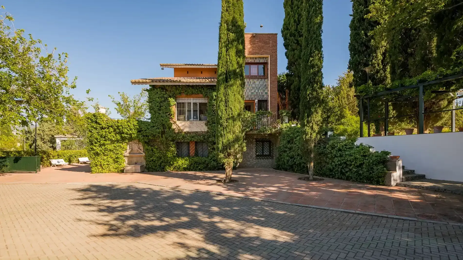 Casa de ladrillo con hiedra y árboles altos junto a un camino pavimentado soleado.